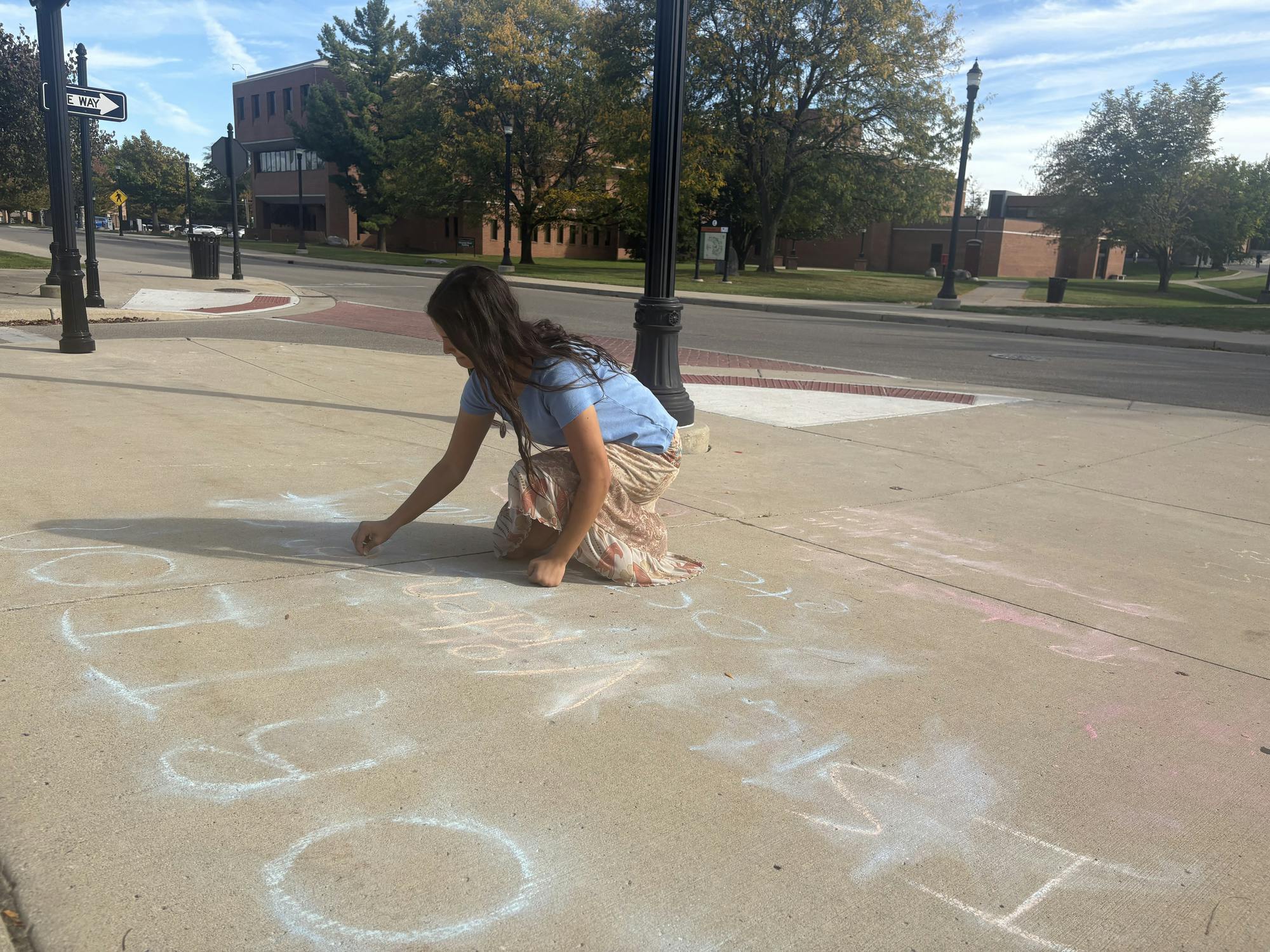 Rosa Skrobola kneels on the sidewalk while writing large letters in chalk.
