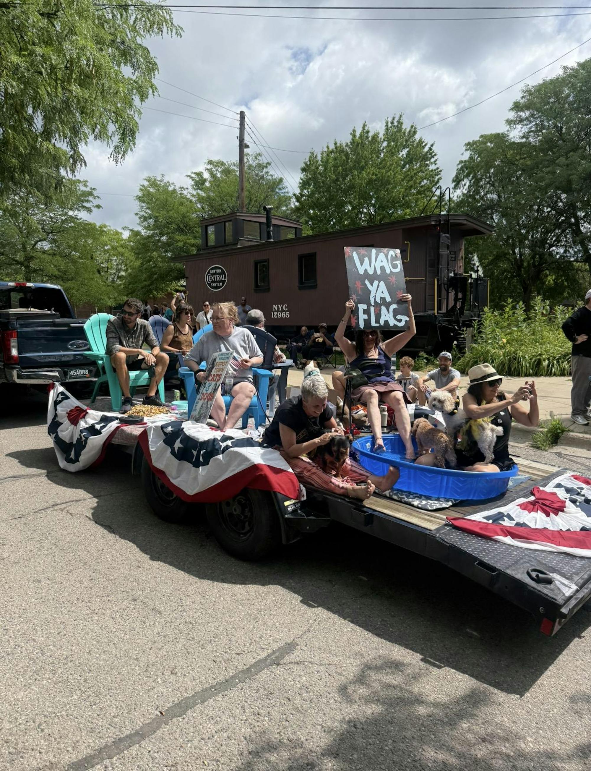 Six people are on top of a Fourth of July parade float decorated with American flag bunting. Several dogs ride along, with a kiddie pool on top. Someone holds a sign saying "Wag ya flag."