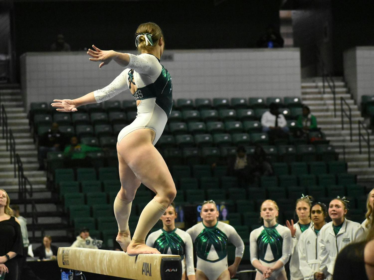 Gymnast Kyrie Lowe poses on the balance beam. Her team watches her in the background.