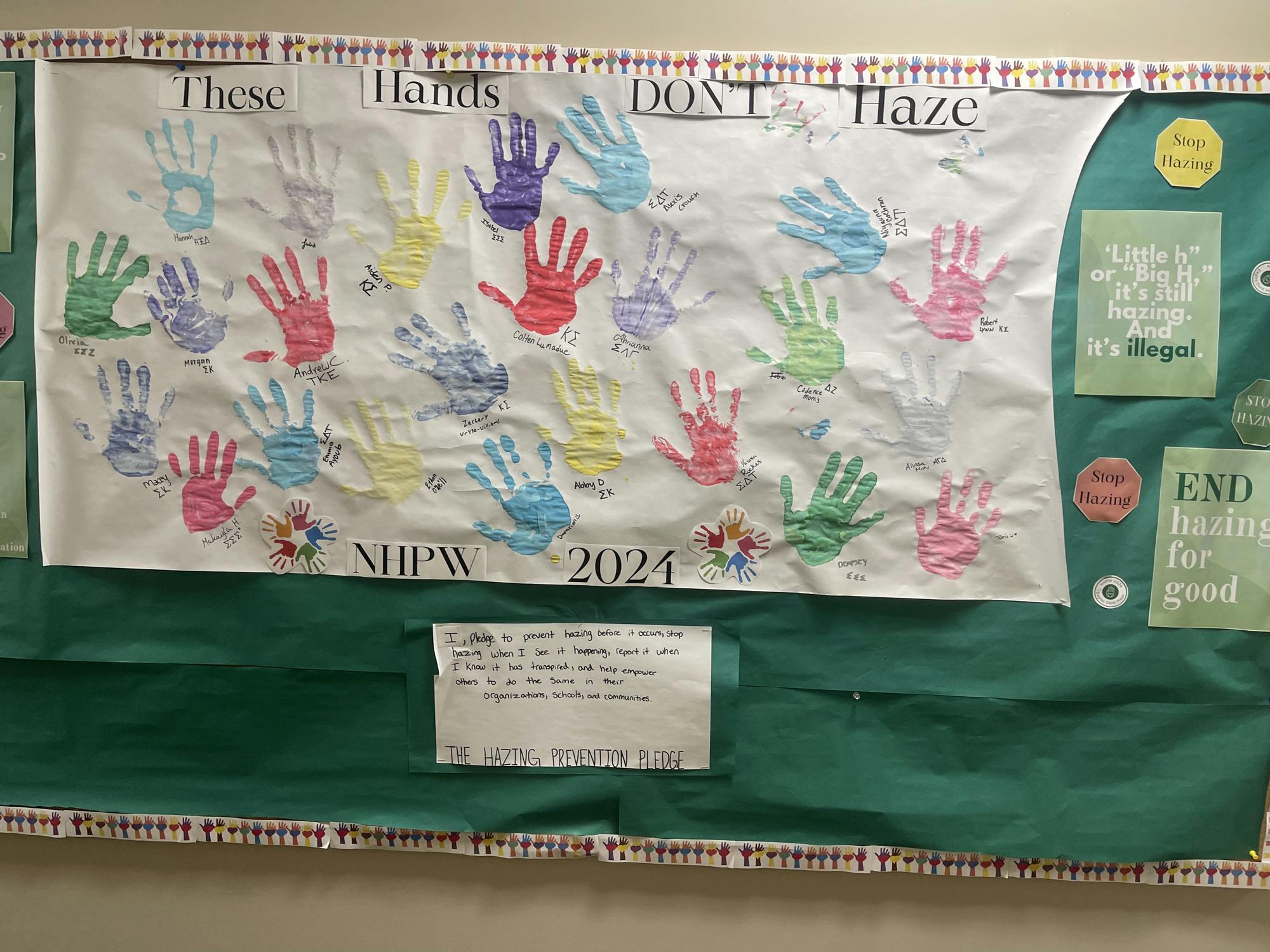 Large paper poster fro 2024 pinned to bulletin board hanging on wall. Poster shows many colorful handprints stamped onto the poster with paint.