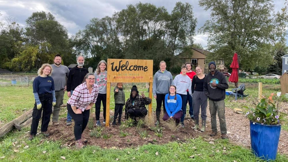 13 people pose for a photo on an outdoor farm. They surround a handmade wooden sign that reads "Welcome, We the People Opportunity Farm."