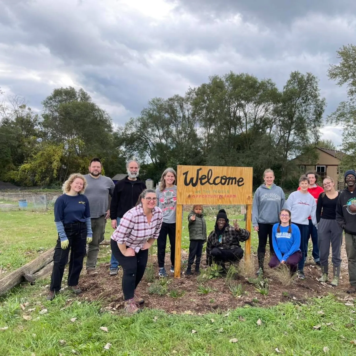 13 people pose for a photo on an outdoor farm. They surround a handmade wooden sign that reads "Welcome, We the People Opportunity Farm."