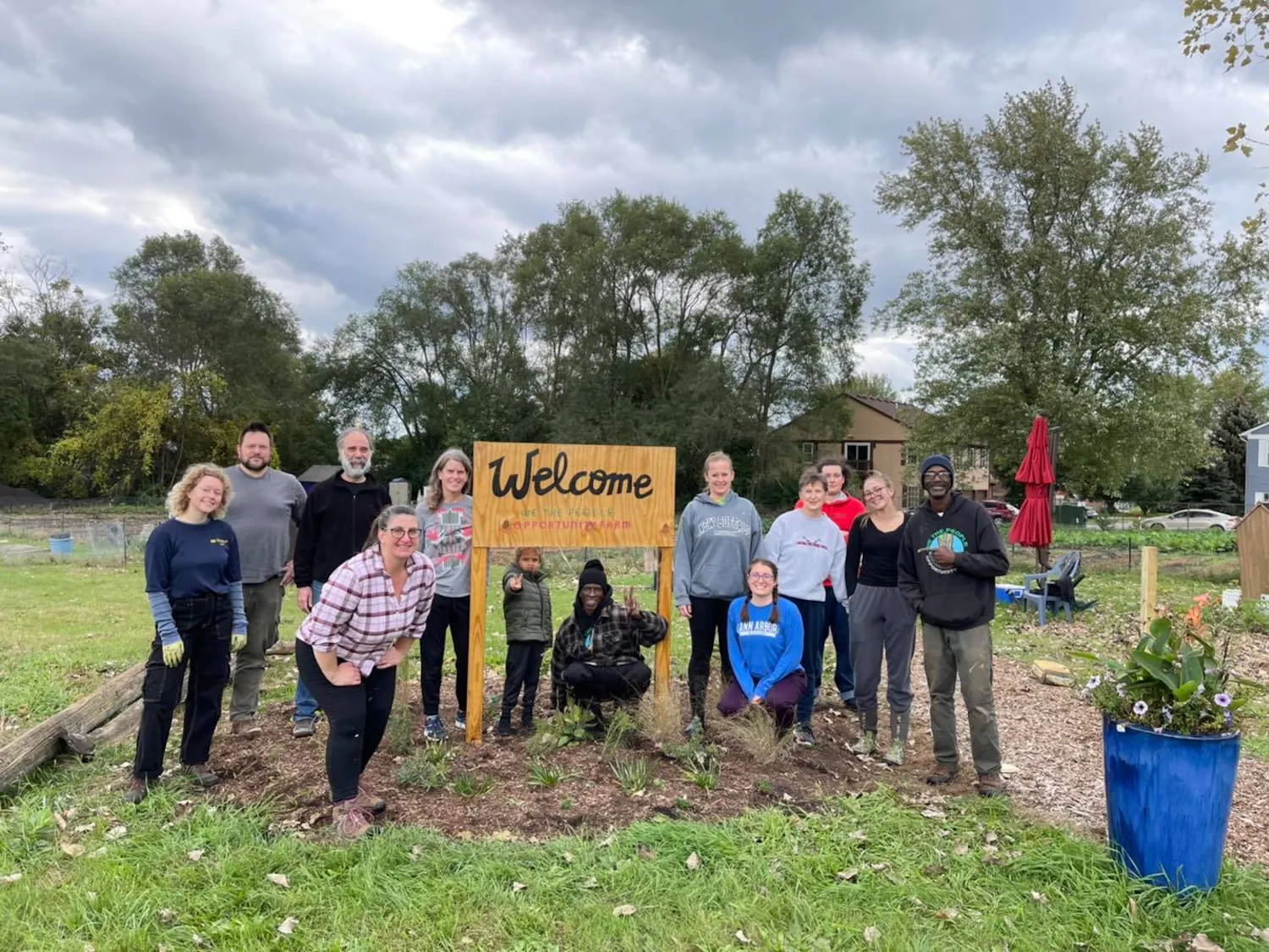 13 people pose for a photo on an outdoor farm. They surround a handmade wooden sign that reads "Welcome, We the People Opportunity Farm."