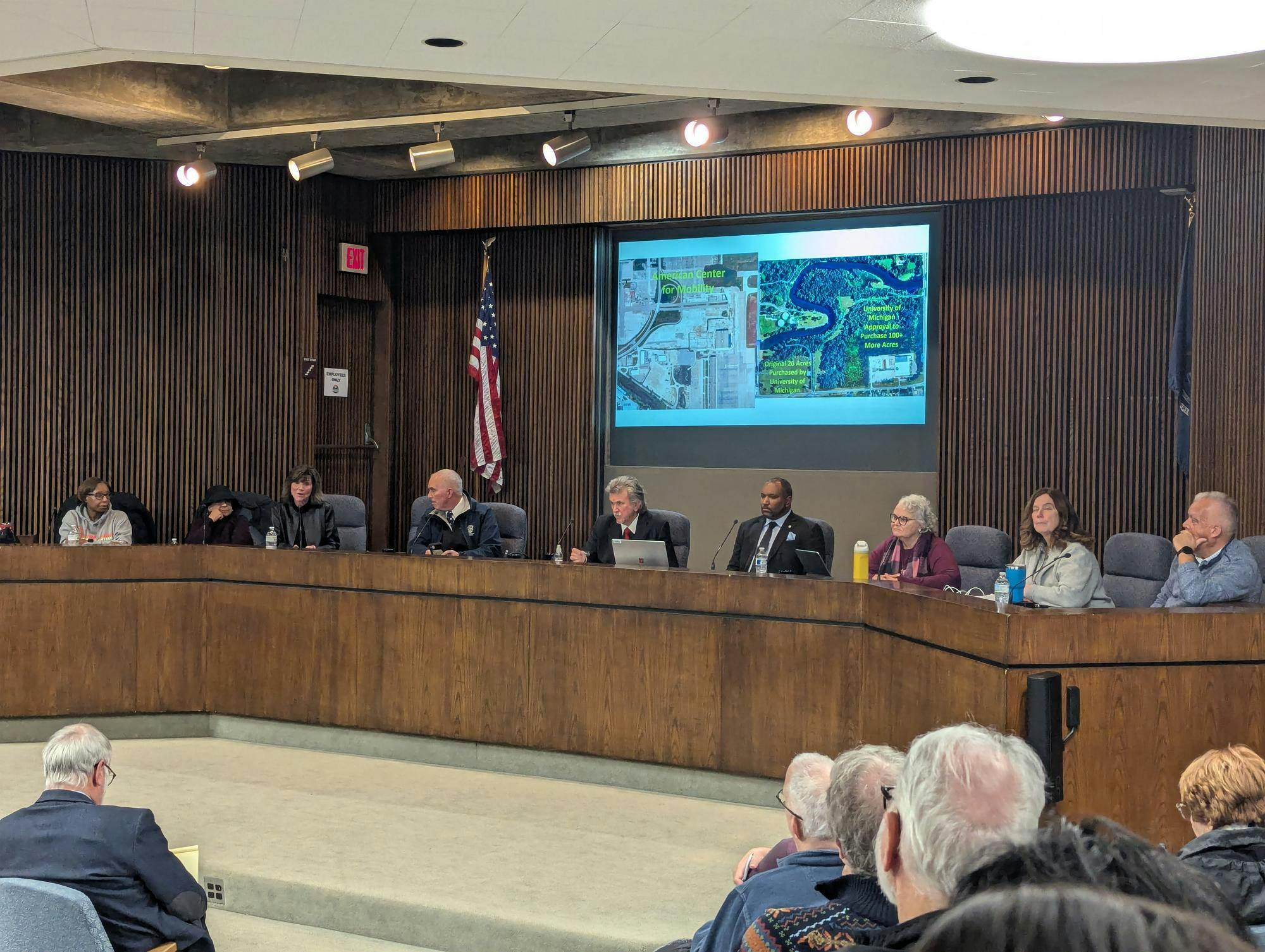 Nine people sit behind a large wooden counter-style desk. Behind them is a projector screen with maps displayed. A few audience members can be seen low in the foreground.