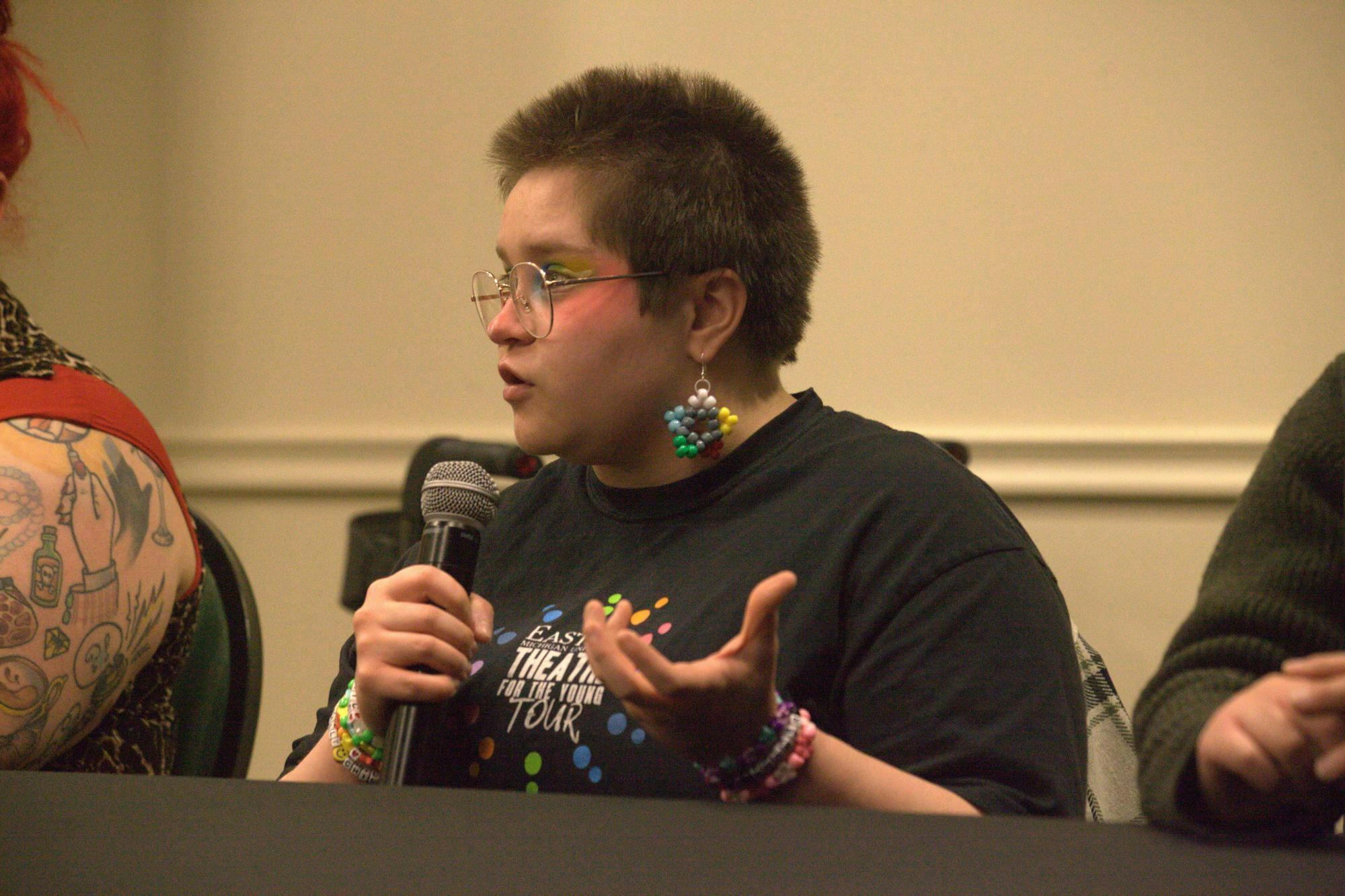 A person speaks while holding a microphone, sitting behind a table indoors. His shirt reads "Eastern Michigan University Theater For the Young Tour."