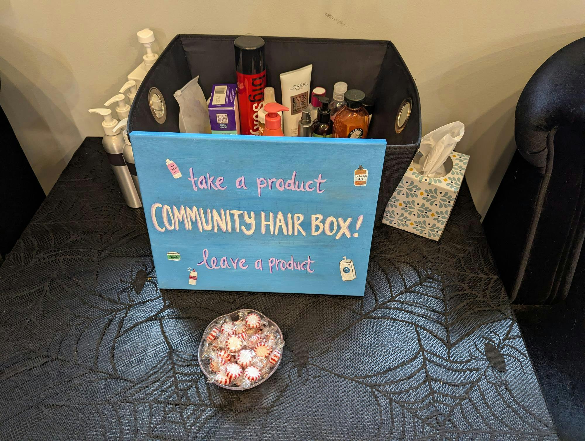 A box of hair products labeled "Take a Product, Leave a Product Community Hair Box" with a blue, hand-painted sign.