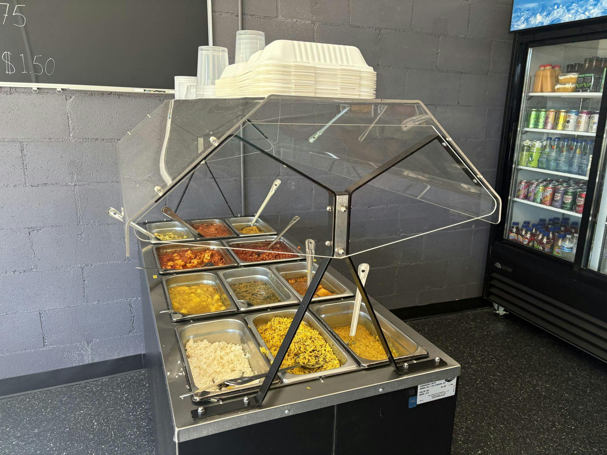 Small, metal buffet counter filled with various Indian cuisine meals, rice, and sauces. A refrigerator displaying food is visible in the background.