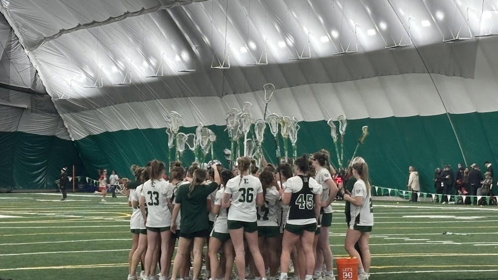 Approximately 20 members of the women's lacrosse team gather on a grass field in an indoor athletics facility.