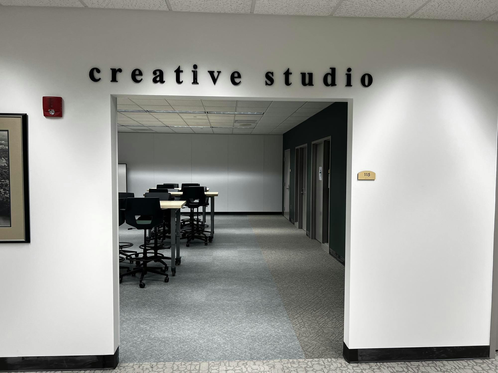 A room with a few high-top tables and chairs. Two podcast studio rooms can be seen on the right wall.