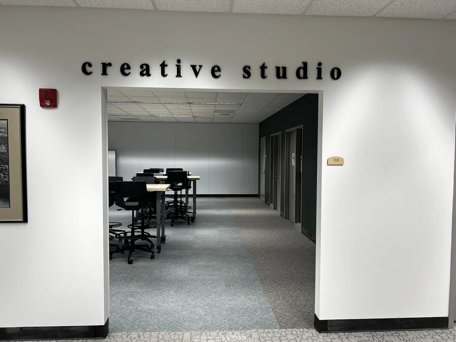 A room with a few high-top tables and chairs. Two podcast studio rooms can be seen on the right wall.