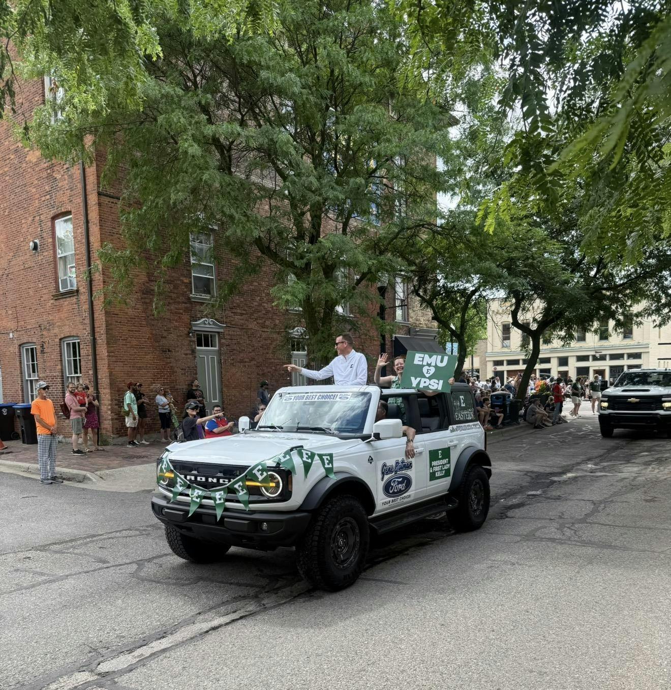 President Kelly stands in a Ford Bronco decorated with Eastern Michigan University logos as part of the parade, while other riders in the float wave at people gathered on the sign. One of the people in the vehicle is holding a sign that says, "EMU [hearts] Ypsi."