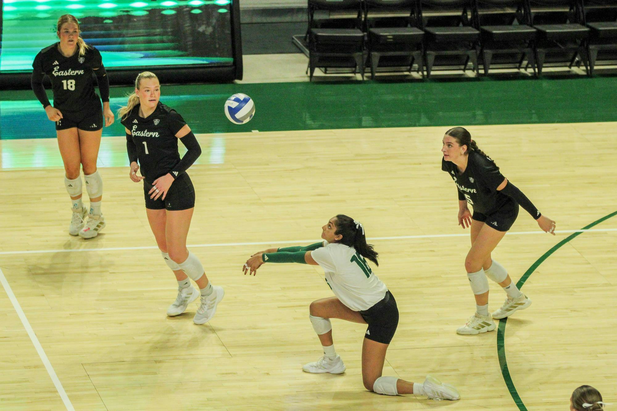 EMU player #15, in a white jersey, bumps the volleyball with her teammates by her on the court, in black jerseys. 