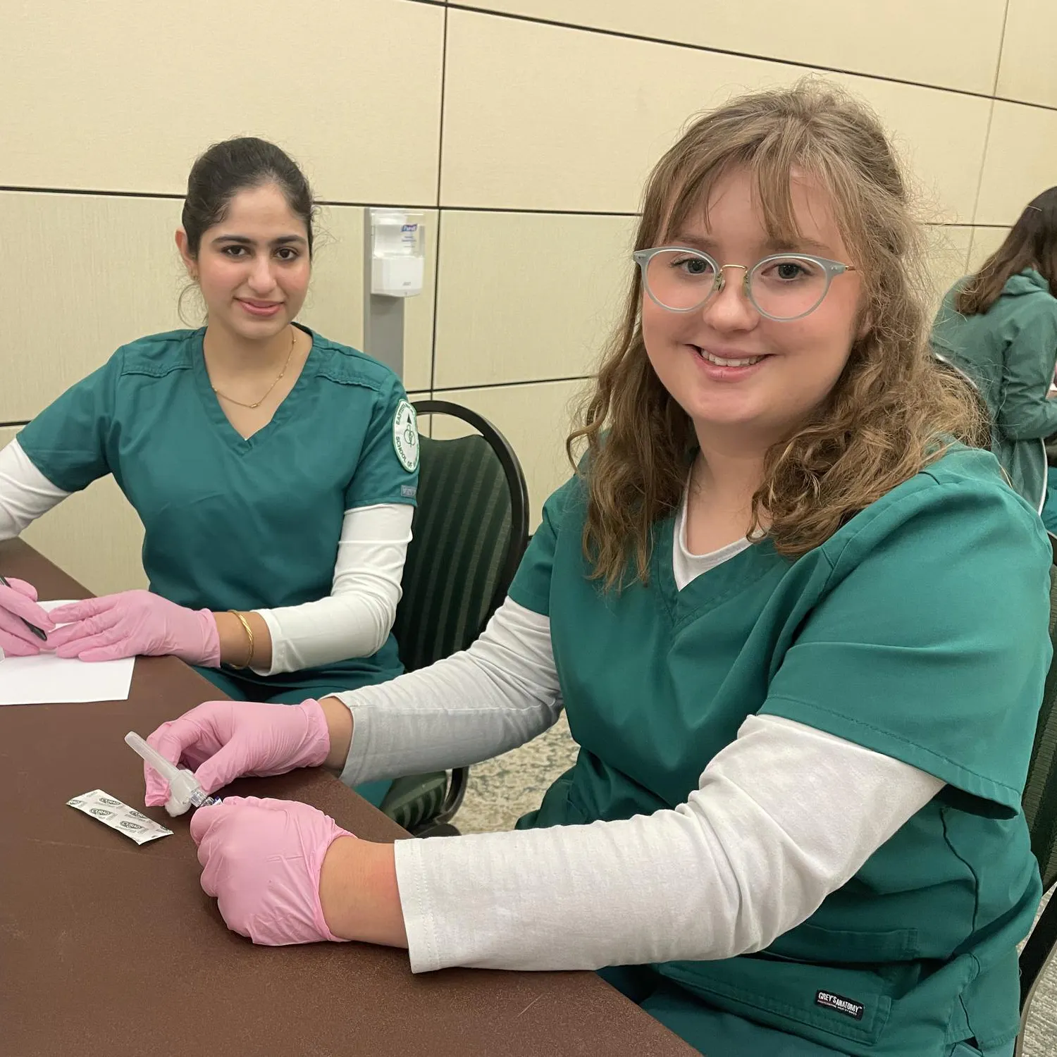 Two people sit at a desk in blue medical scrubs and pink gloves. One of them holds a syringe, and another holds a pen.