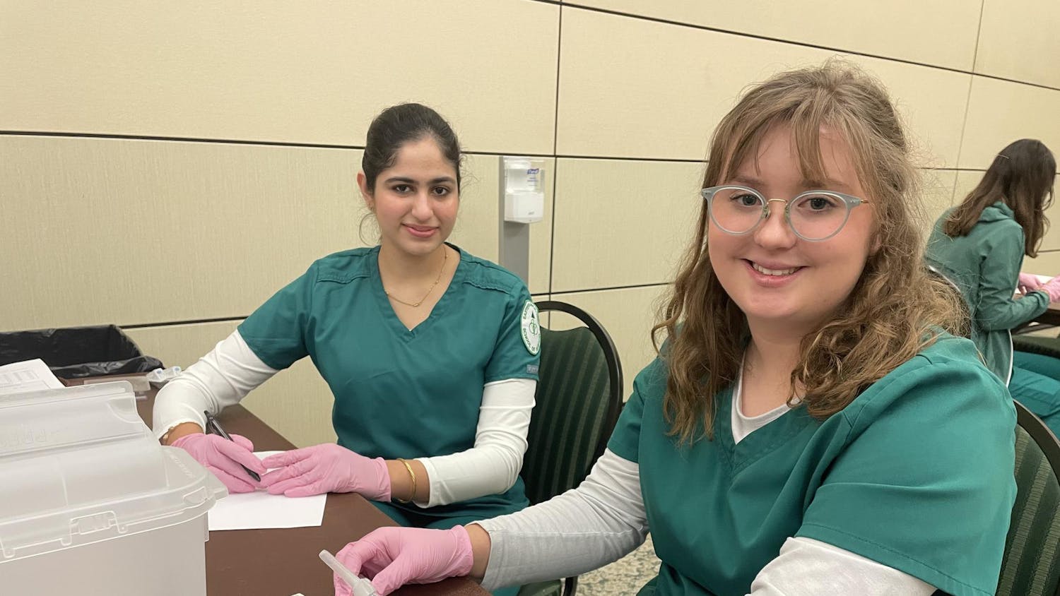 Two people sit at a desk in blue medical scrubs and pink gloves. One of them holds a syringe, and another holds a pen.