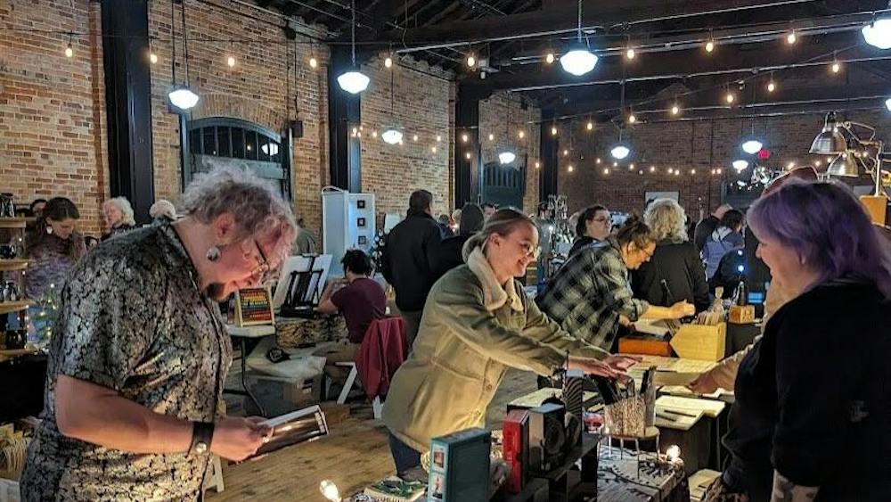 A group of people admire items on tables sold by vendors in a brick-walled building.