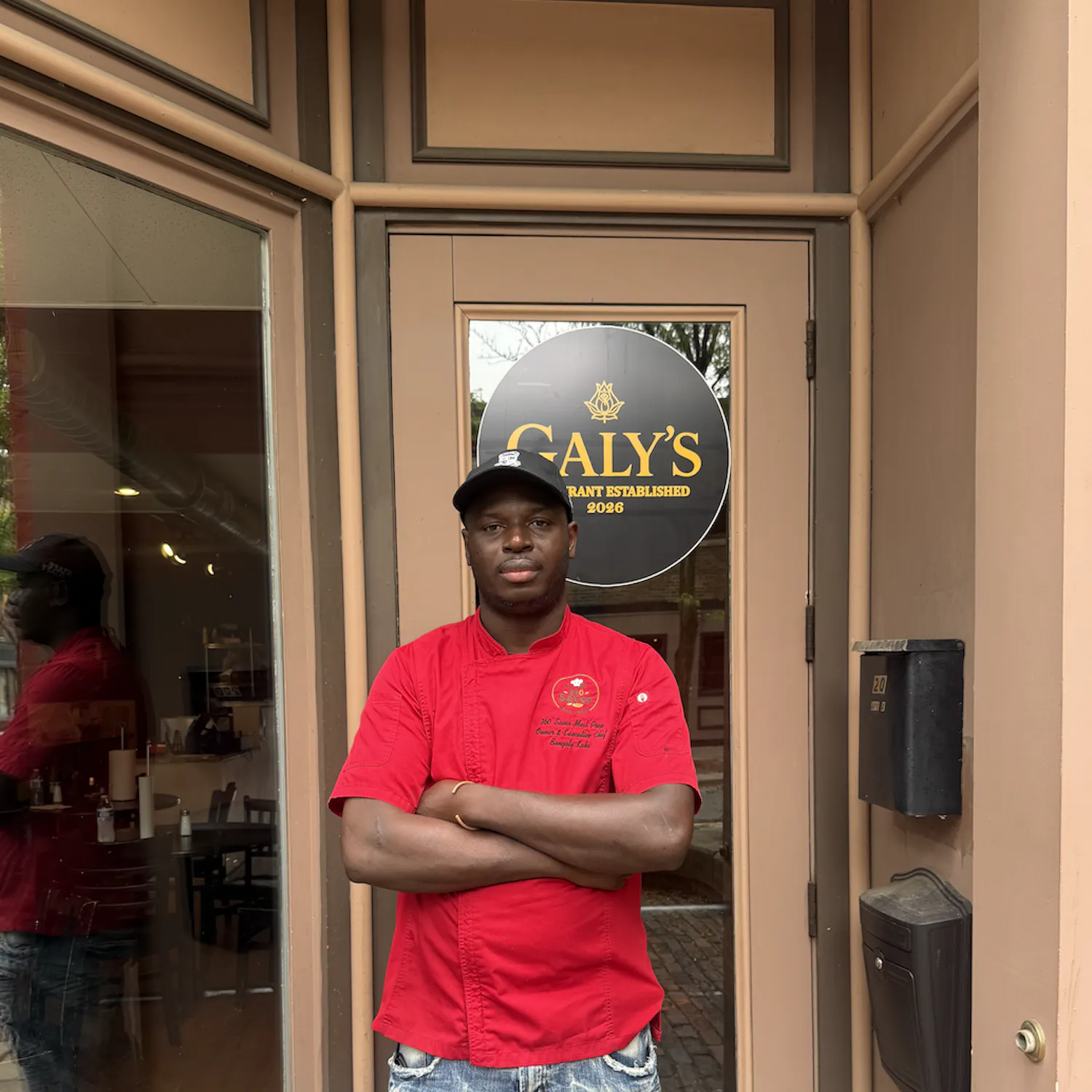 A man in a red chef's coat stands in front of his restaurant.