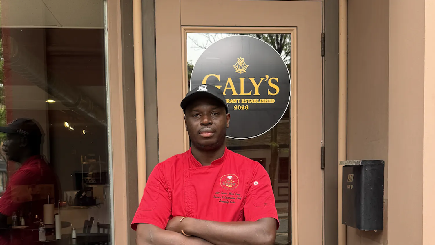 A man in a red chef's coat stands in front of his restaurant.