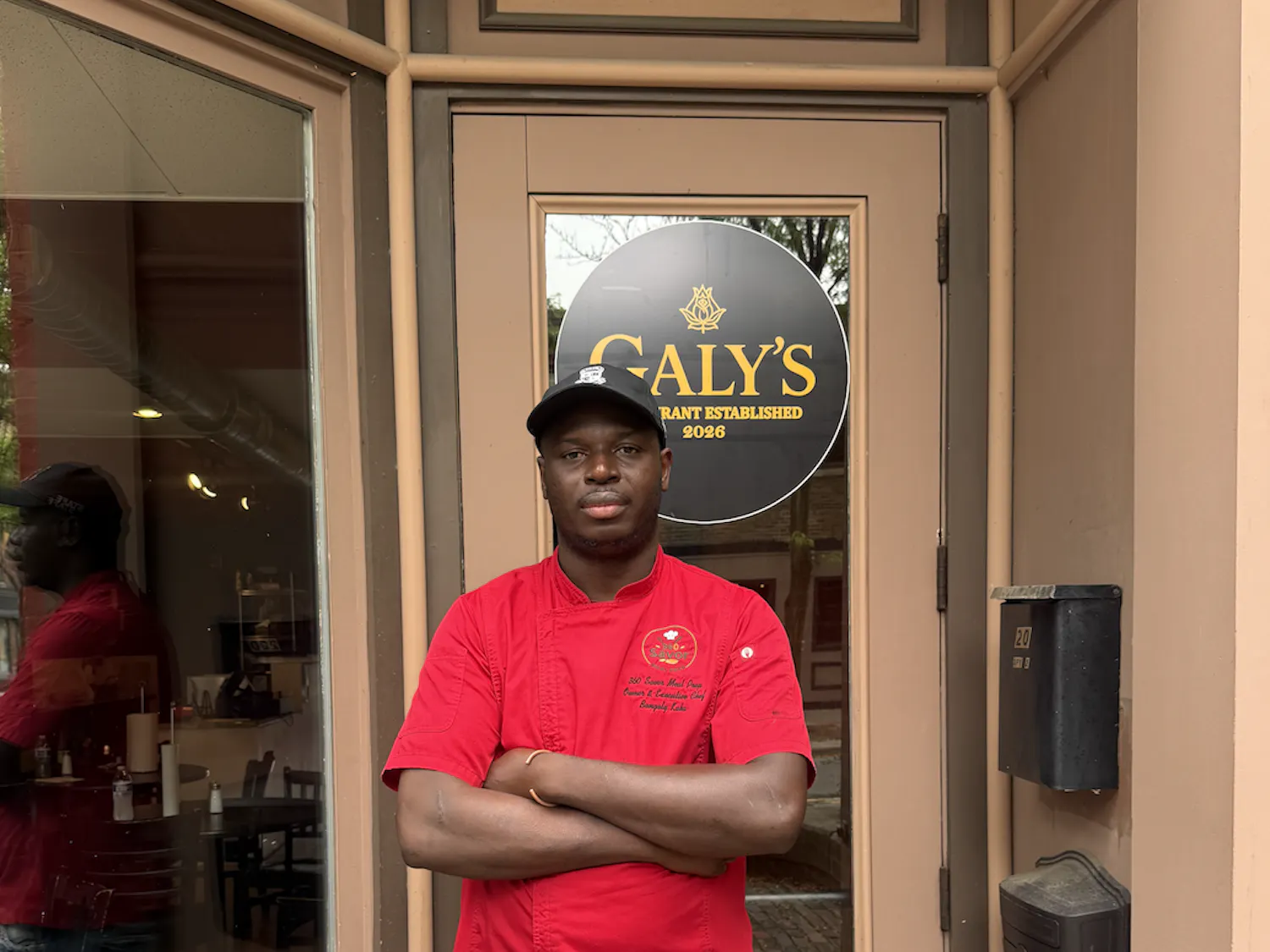 A man in a red chef's coat stands in front of his restaurant.