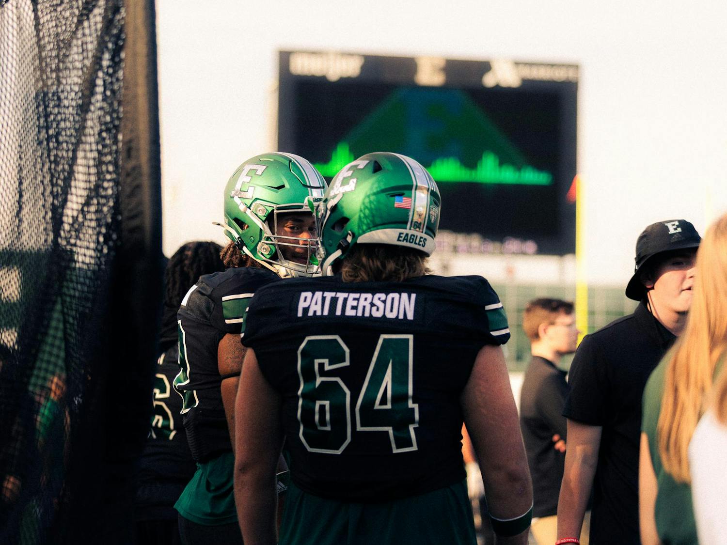 Two Eastern Michigan offensive linemen, both decked out in green and black wearing green helmets with a stylized grey "E", converse during the team's Homecoming win against ULL.