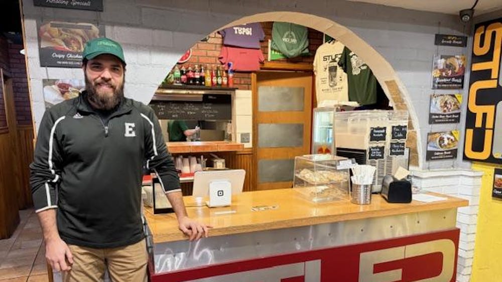 Joel Hamami stands in front of the ordering counter, which says, "STUFD" on the front. In the background there are branded T-shirts and menu item photos.