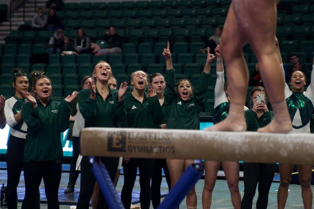 A group of EMU gymnasts wearing green and white cheer and clap for their teammate on the beam.