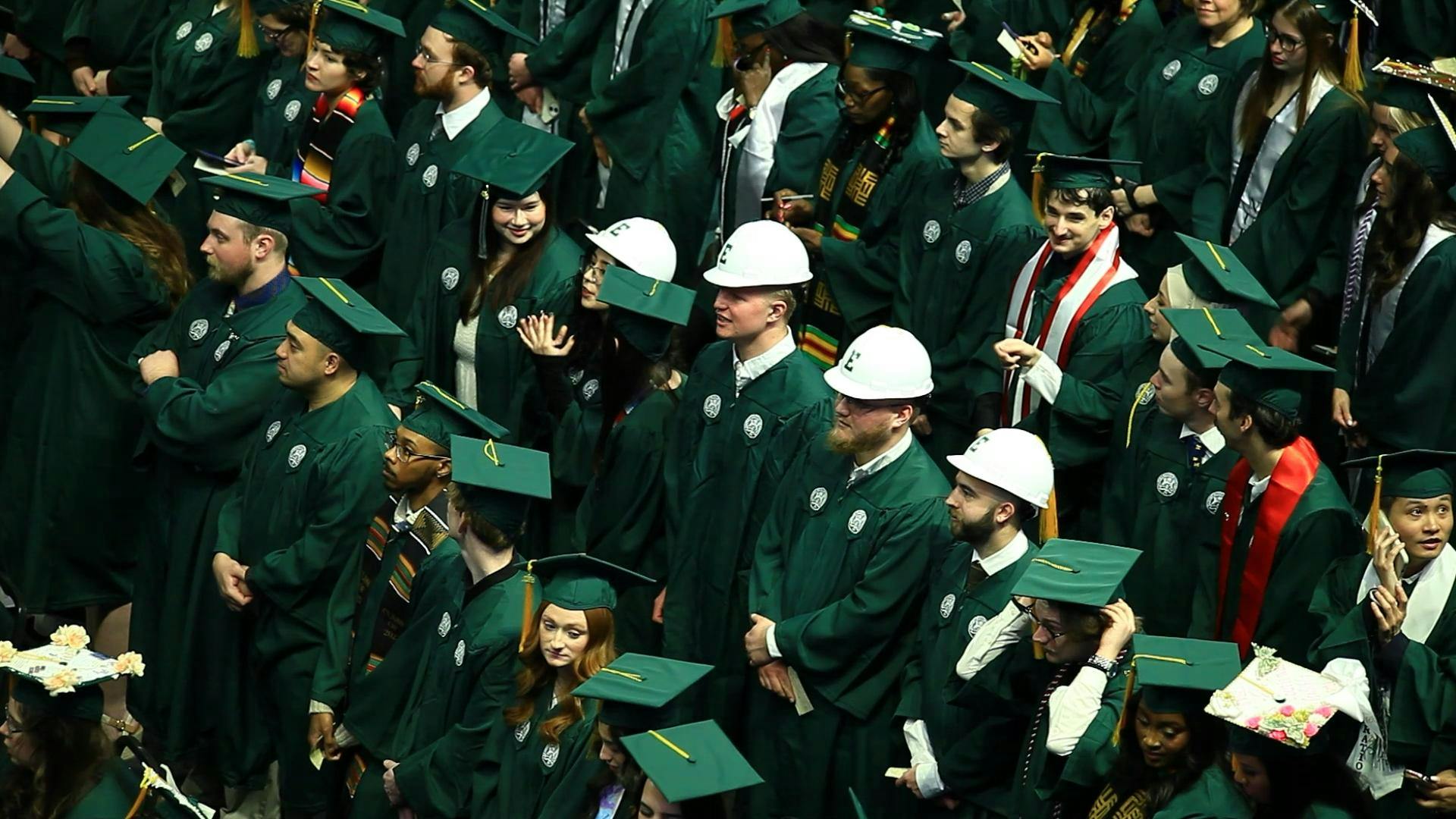 A crowd of students in green graduation gowns and caps stand in rows, with another row in the middle wearing white hard hats with their graduation uniform. 