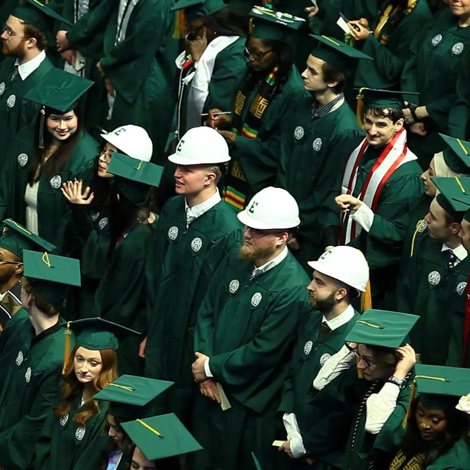 A crowd of students in green graduation gowns and caps stand in rows, with another row in the middle wearing white hard hats with their graduation uniform.