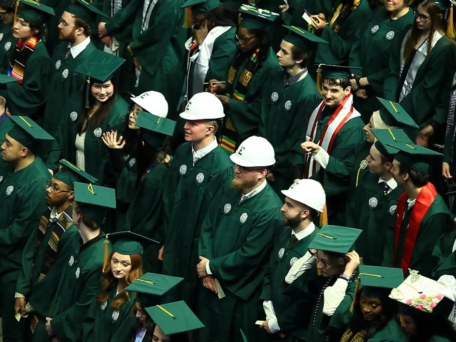 A crowd of students in green graduation gowns and caps stand in rows, with another row in the middle wearing white hard hats with their graduation uniform.
