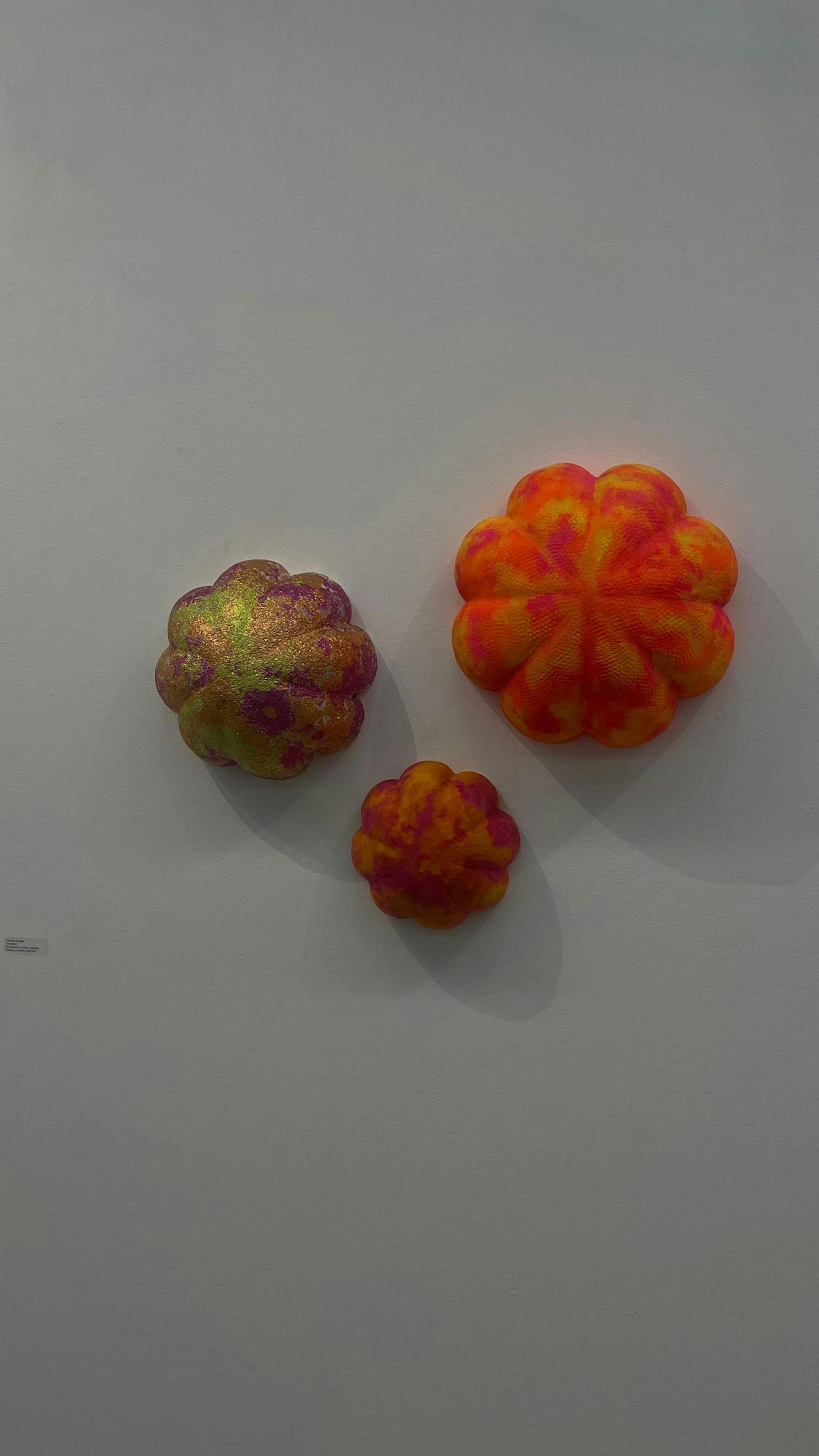 Three flower-shaped art pieces hang on display against a blank wall. They are yellow, orange, and pink, with the leftmost being shiny and the others in neon colors. Each has a rough texture.