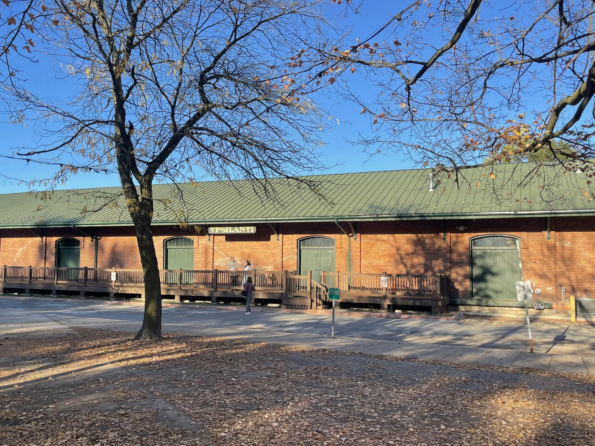 The exterior of the Ypsilanti Freighthouse, a long, slender building made of red bricks with a dark green roof. A sign hanging from the building's roof says "Ypsilanti" in gold-colored letters. The sky overheard is blue, and there is a tree in the foreground of the photo that has lost most of its autumn leaves, which are now on the ground around it.
