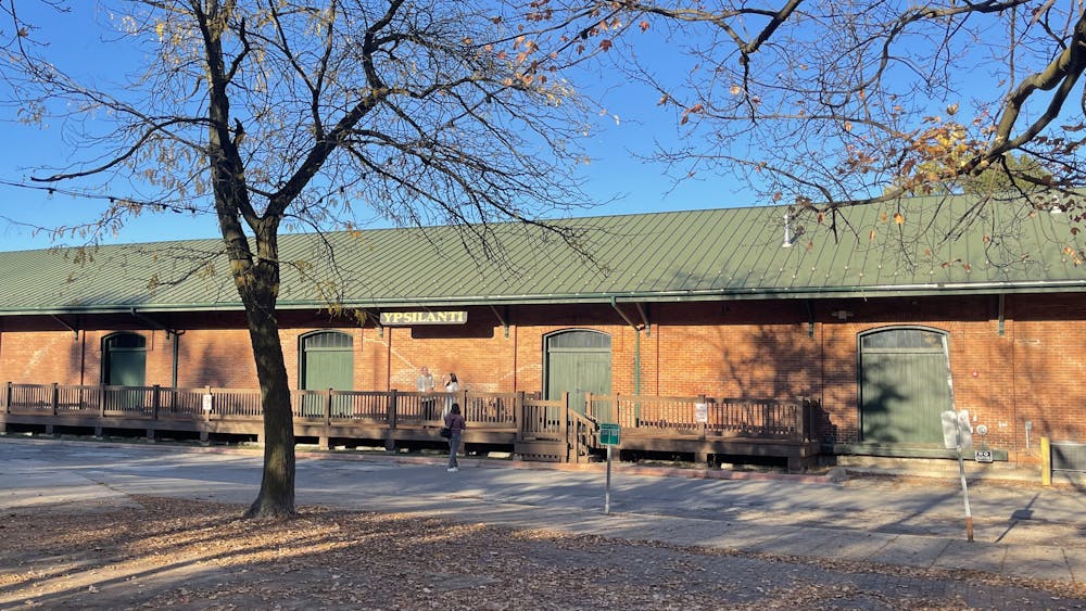 The exterior of the Ypsilanti Freighthouse, a long, slender building made of red bricks with a dark green roof. A sign hanging from the building's roof says "Ypsilanti" in gold-colored letters. The sky overheard is blue, and there is a tree in the foreground of the photo that has lost most of its autumn leaves, which are now on the ground around it.
