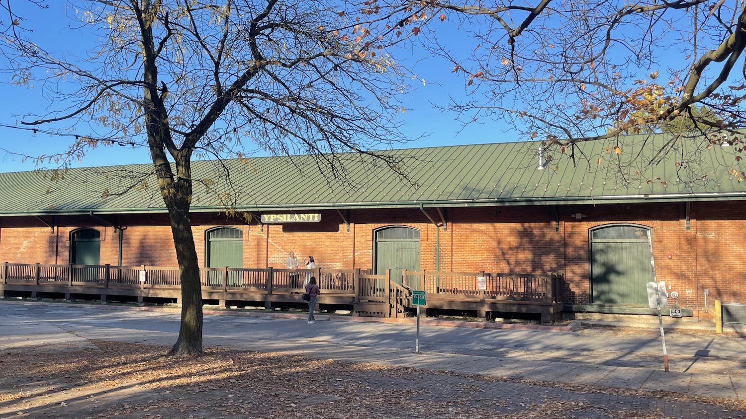 The exterior of the Ypsilanti Freighthouse, a long, slender building made of red bricks with a dark green roof. A sign hanging from the building's roof says "Ypsilanti" in gold-colored letters. The sky overheard is blue, and there is a tree in the foreground of the photo that has lost most of its autumn leaves, which are now on the ground around it.
