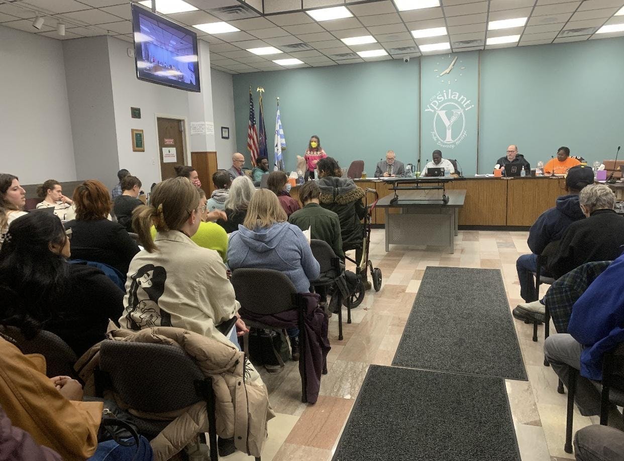 The audience seating during the City Council meeting is nearly full with community members. The Ypsilanti City Council members sit behind a large shared desk at the back of the room.