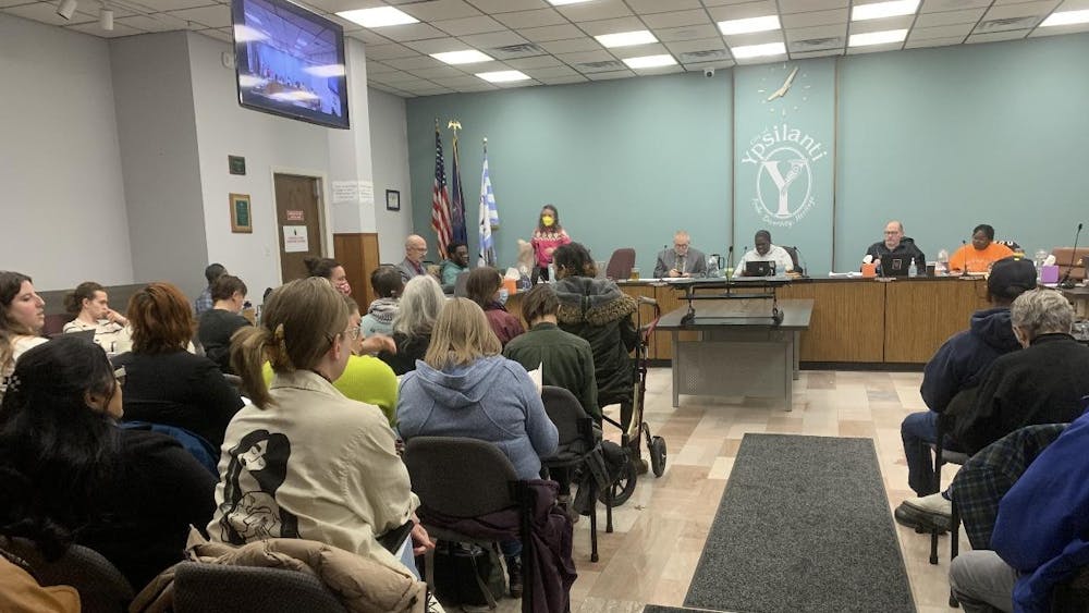 The audience seating during the City Council meeting is nearly full with community members. The Ypsilanti City Council members sit behind a large shared desk at the back of the room.