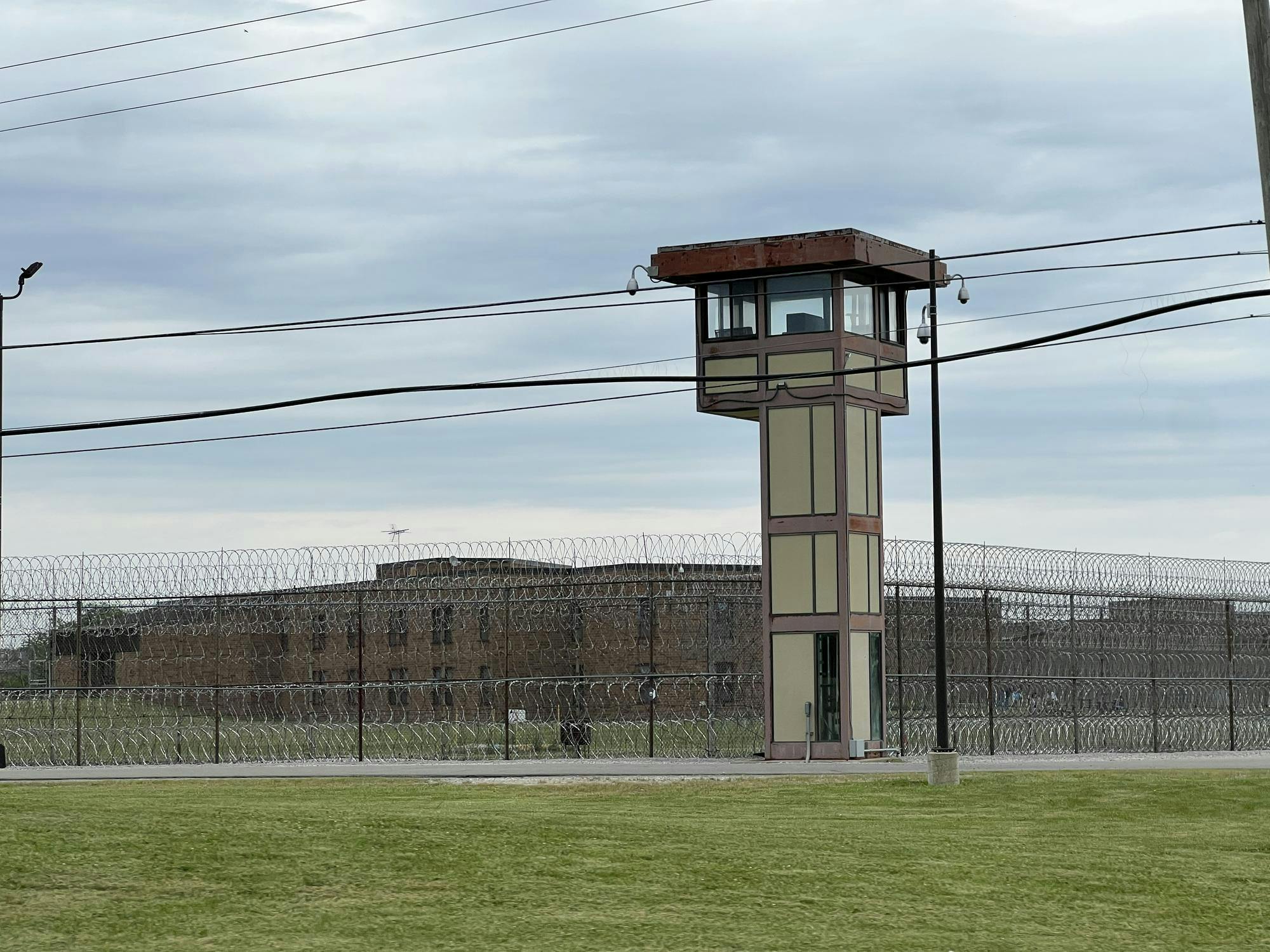 A watchtower surrounded by multiple security cameras stands in front of a barbed wire fence at the edge of the Women's Huron Valley Correctional Facility yard.