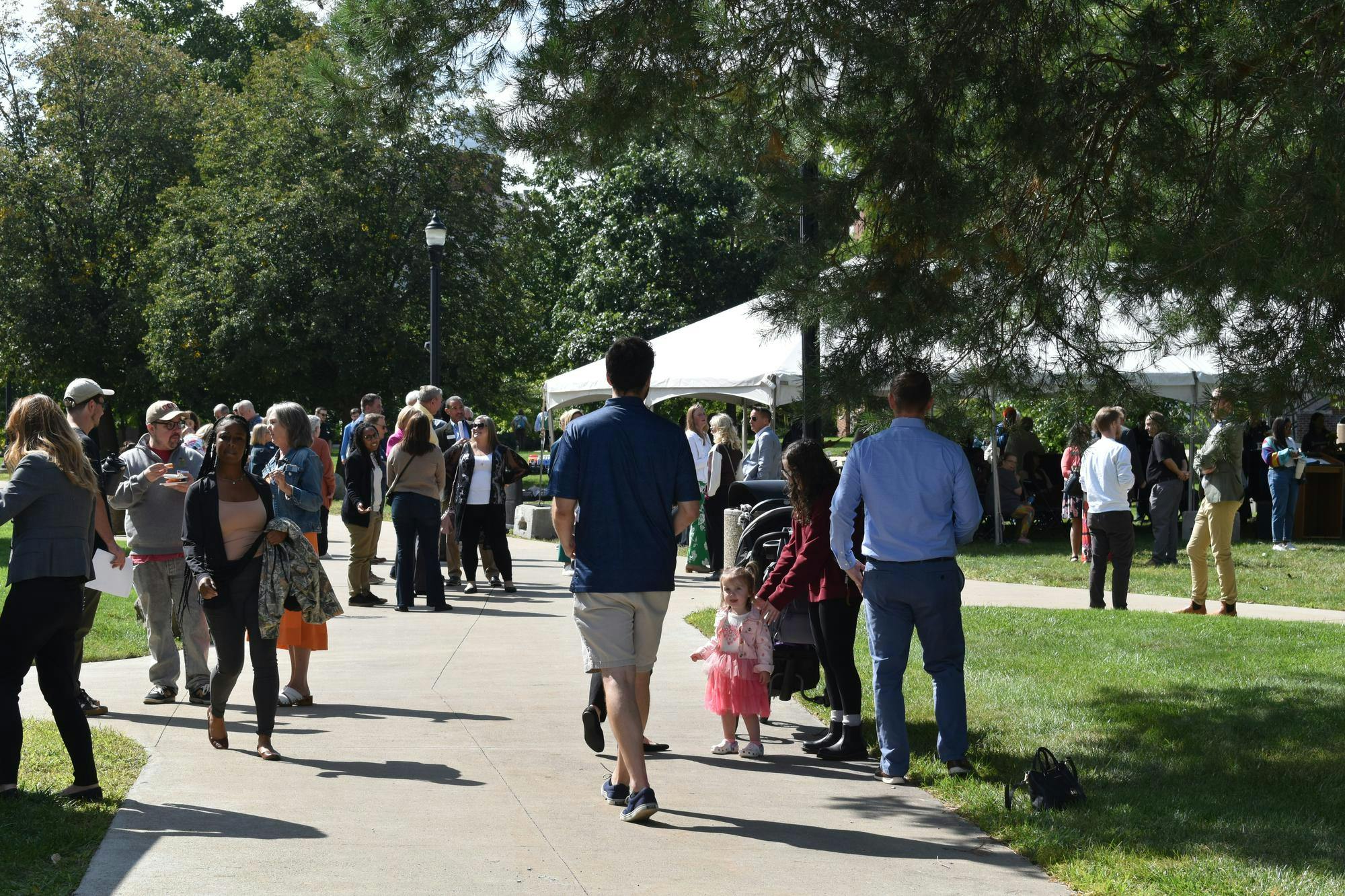 Event attendees walk and gather on the sidewalk and lawn outside of a white canopied tent that is pitched in the background.
