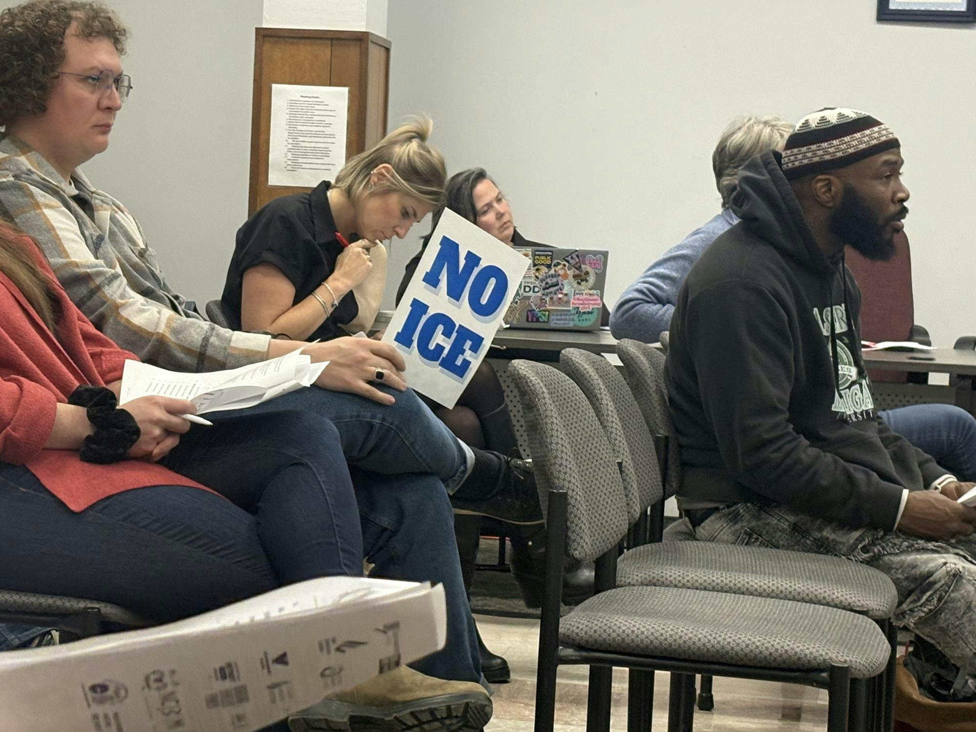 Multiple people sit in rows of chairs and at a table perpendicular to the chairs at an Ypsilanti City Council meeting, with one person holding a sign that says, "No ICE."
