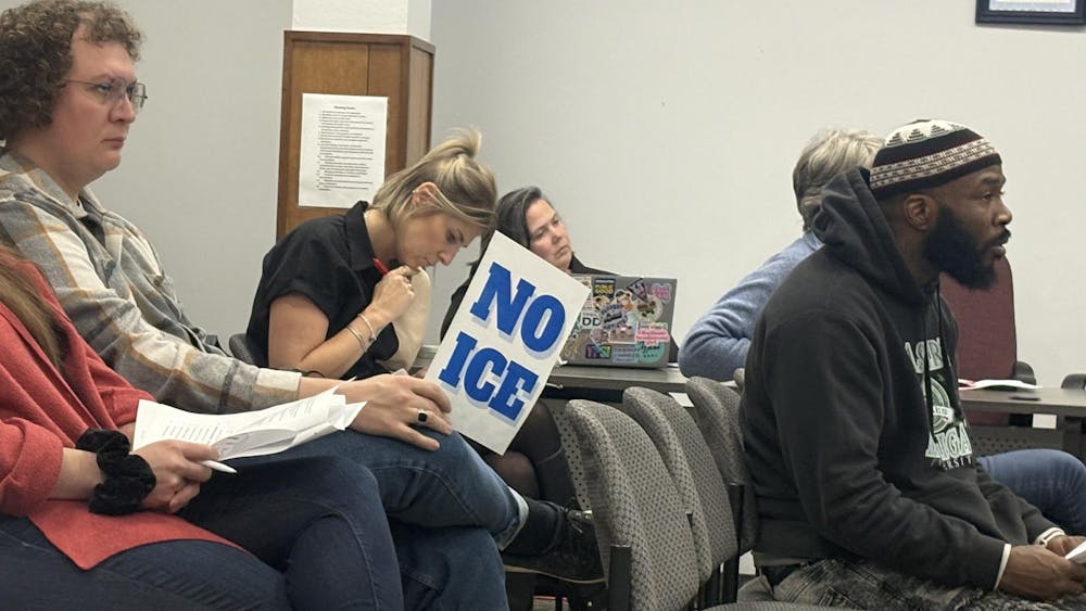 Multiple people sit in rows of chairs and at a table perpendicular to the chairs at an Ypsilanti City Council meeting, with one person holding a sign that says, "No ICE."