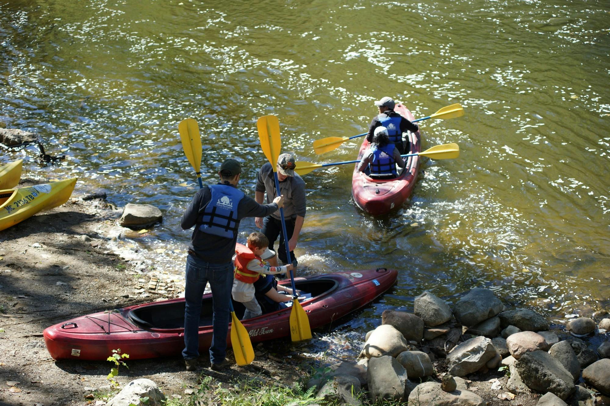 Two red kayaks with yellow paddles enter the river. The leftmost remains halfway on the bank as two individuals help a child into the kayak.