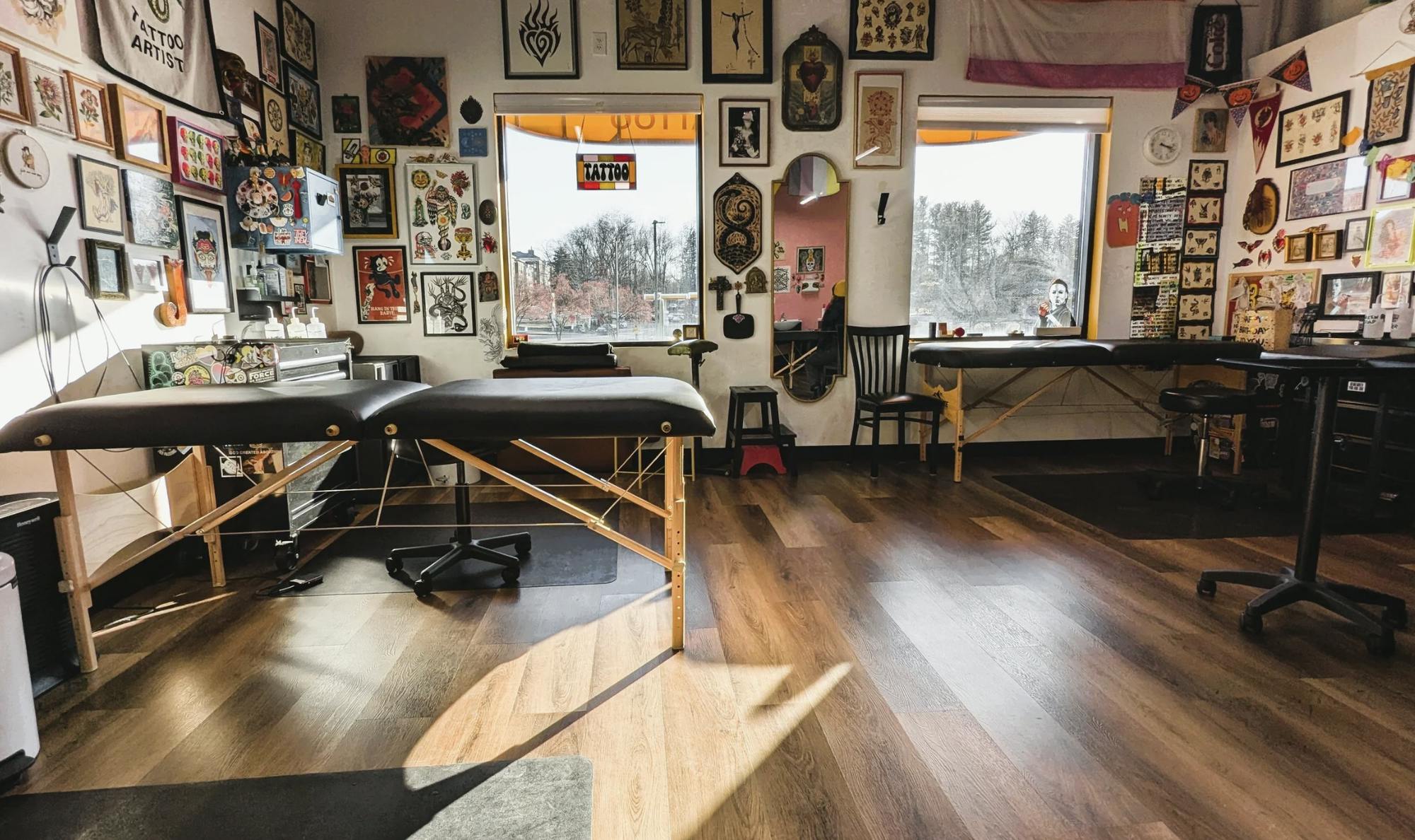 Room with natural lighting streaming through two windows. Two tattoo benches are set up and an eclectic array of decorations cover the wall.
