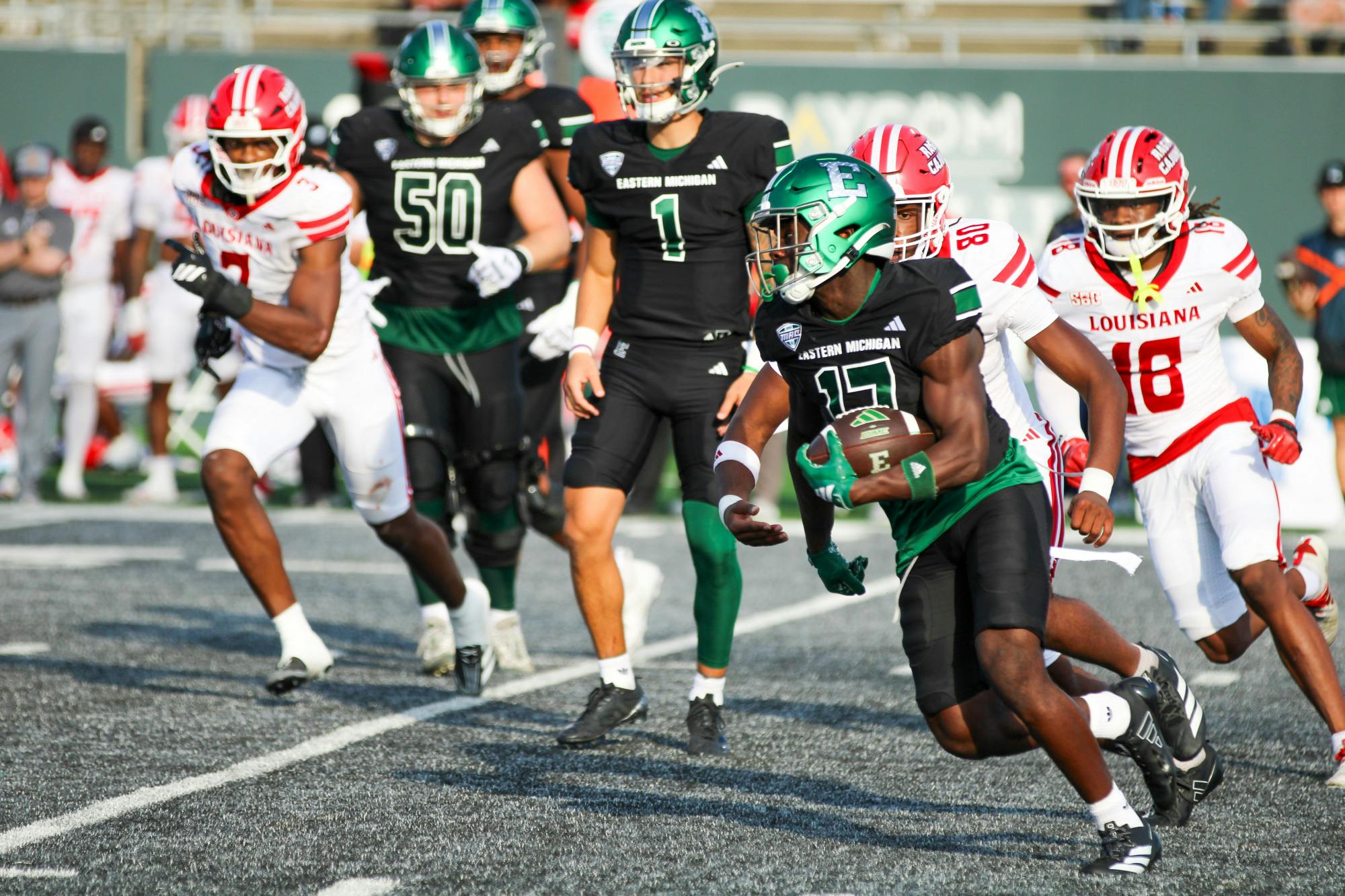 An Eastern Michigan football player dressed in black and green carries the ball with three ULL defenders dressed in red and white in tow. 