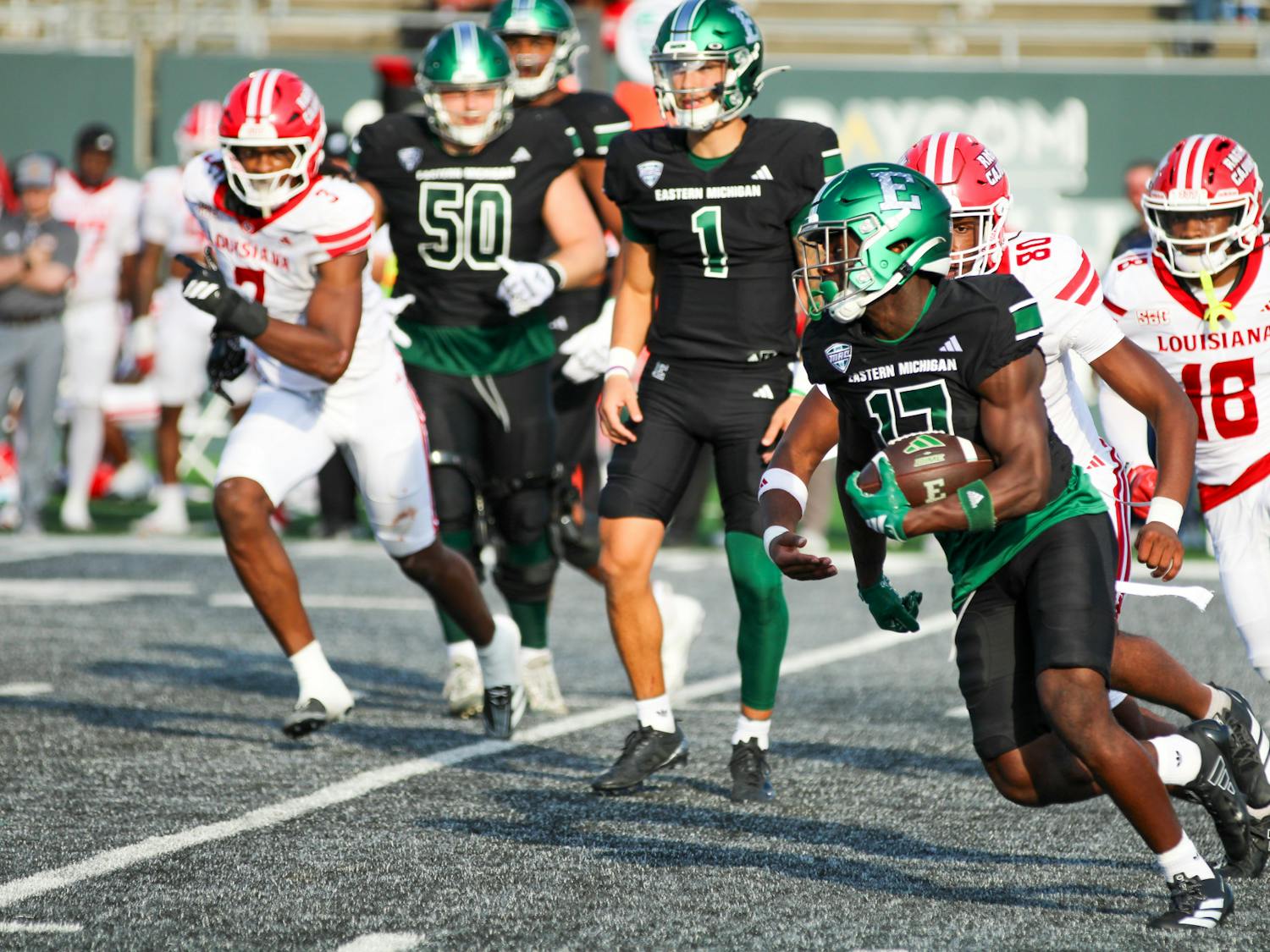An Eastern Michigan football player dressed in black and green carries the ball with three ULL defenders dressed in red and white in tow.