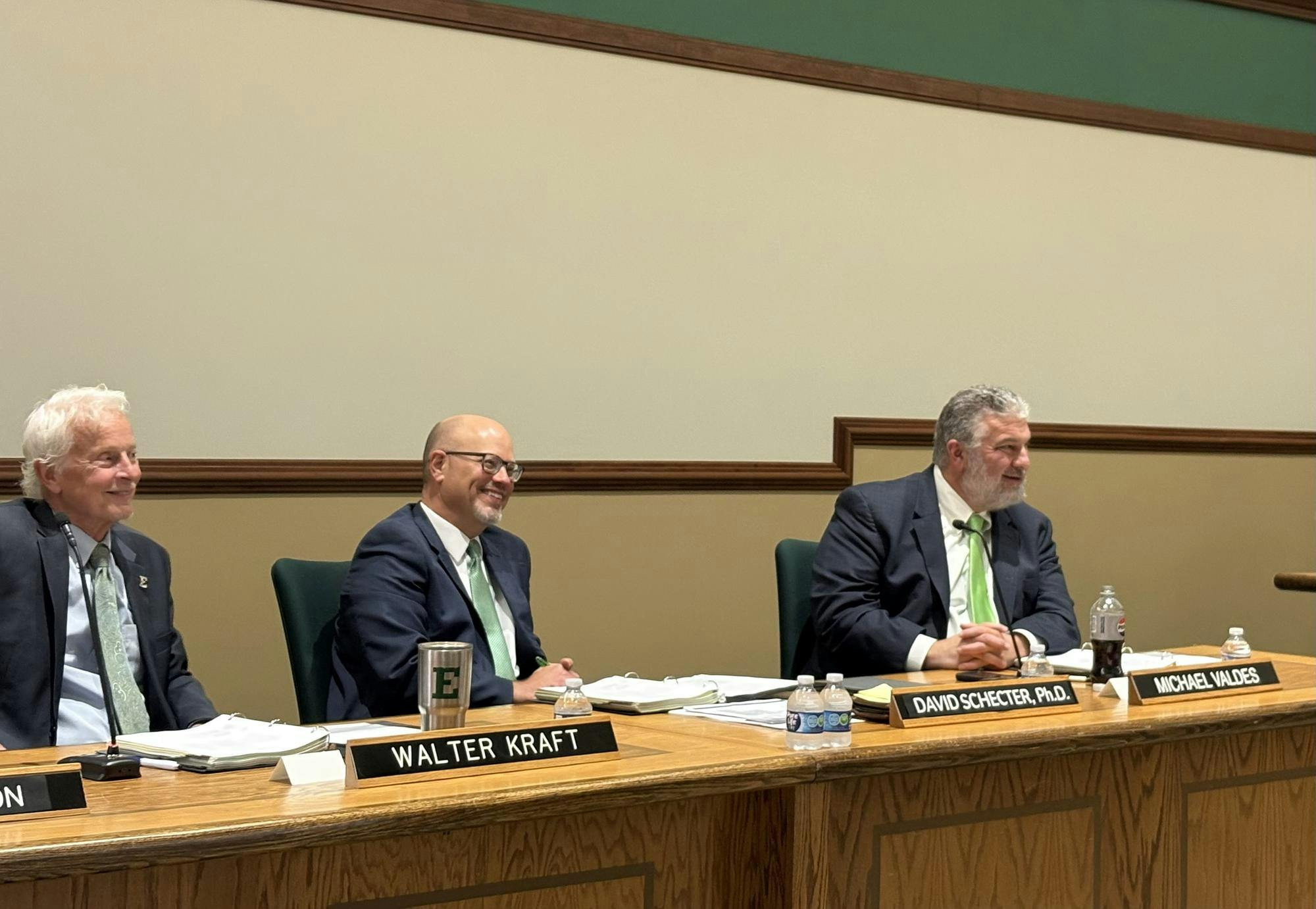 3 men sit behind a long, shared, wooden desk, each of them smiling. A name plate rests on the desk in front of each of them.