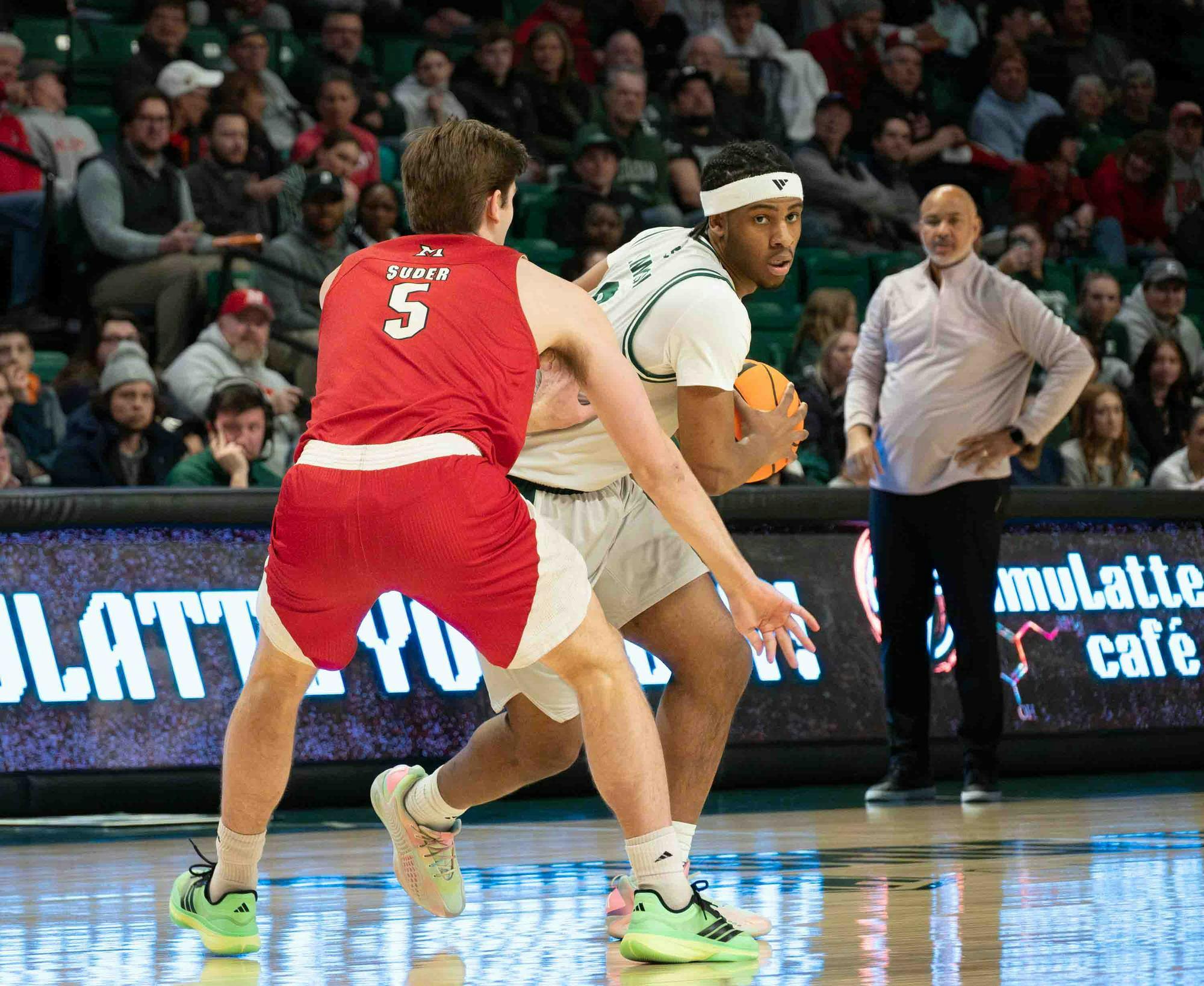 Basketball player from opposing team stands in front of Gregory Lawson II with back facing the camera. Lawson holds the basketball and leans around the player.