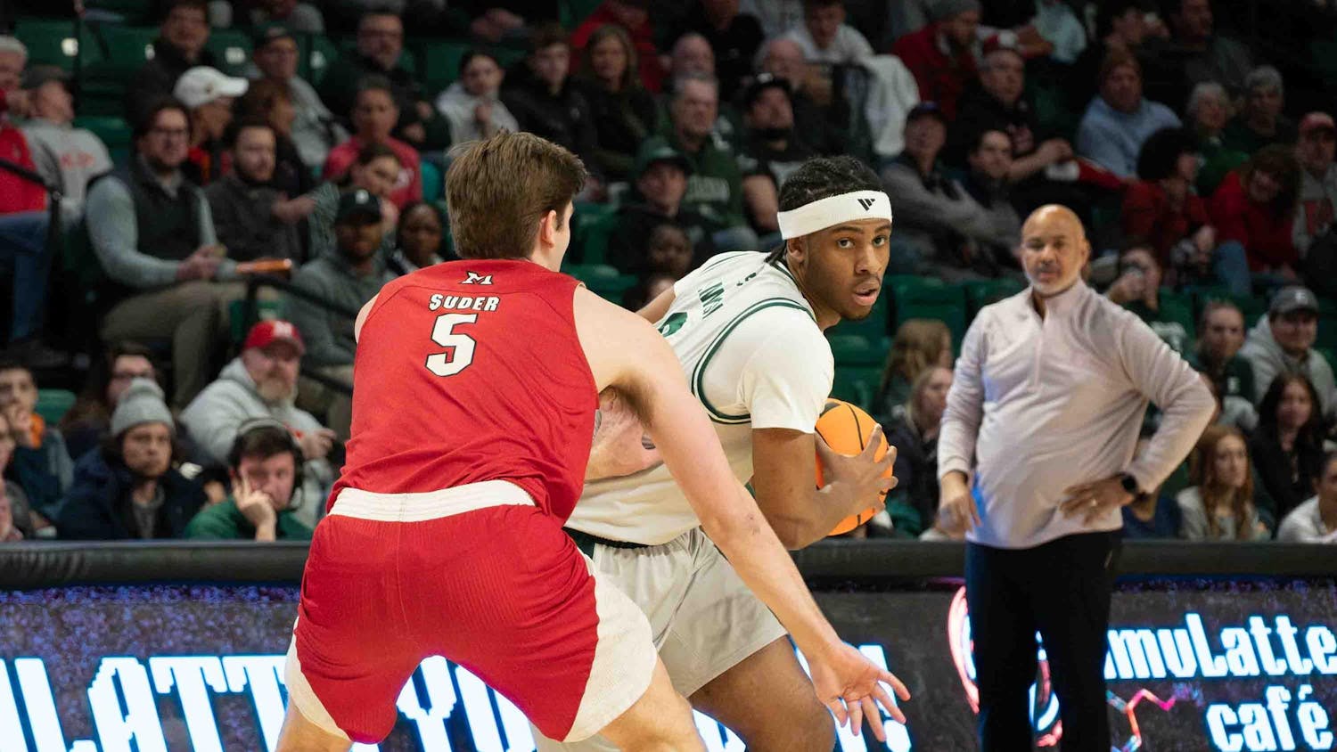 Basketball player from opposing team stands in front of Gregory Lawson II with back facing the camera. Lawson holds the basketball and leans around the player.