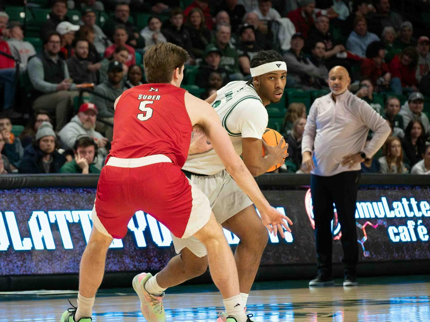 Basketball player from opposing team stands in front of Gregory Lawson II with back facing the camera. Lawson holds the basketball and leans around the player.