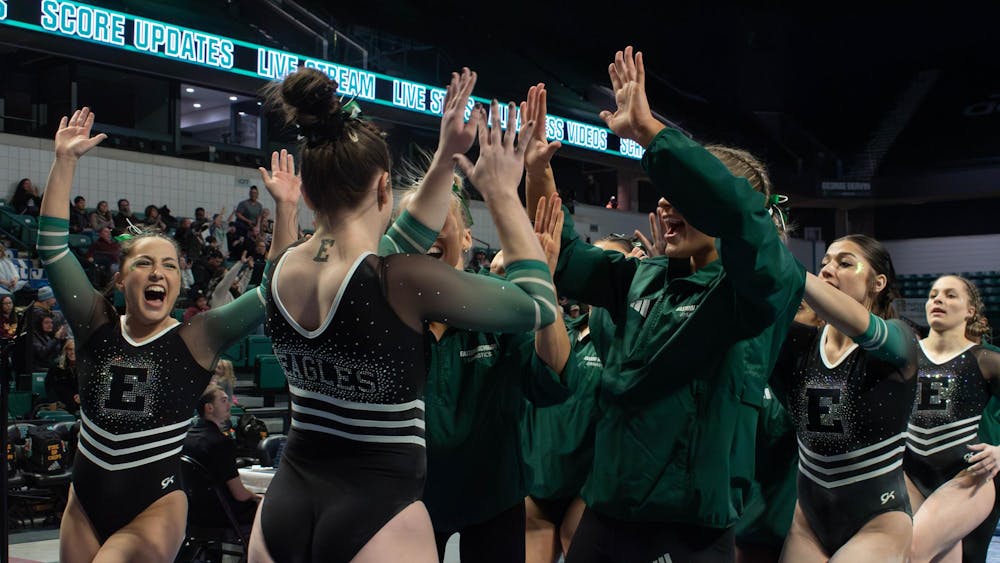 Several college-aged gymnasts wearing black and green Eastern Michigan University leotards energetically high-five one another and smile.