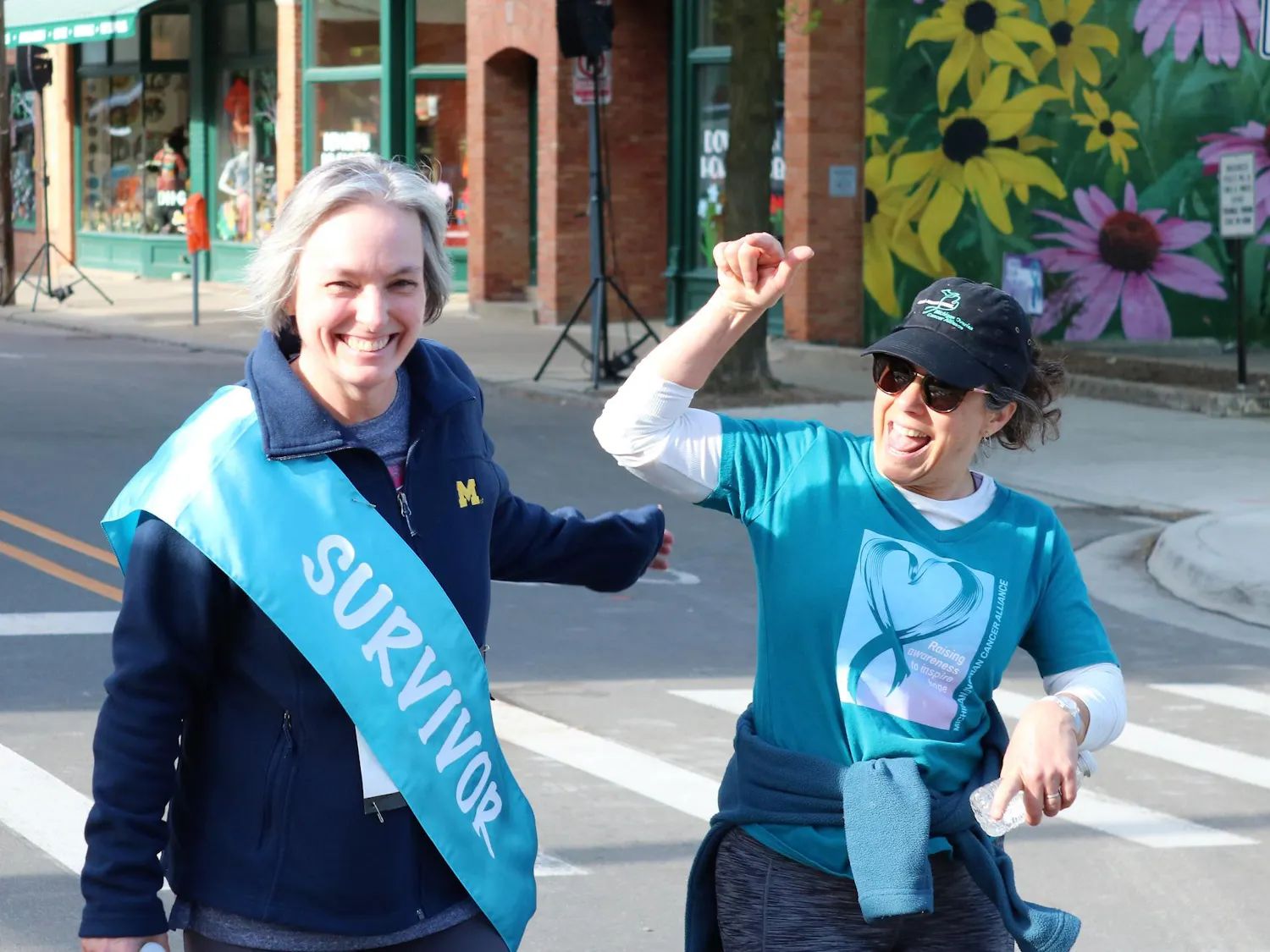 Two women celebrate and smile while wearing blue shirts for the event, with the leftmost wearing a teal survivor ribbon as a sash across her chest.