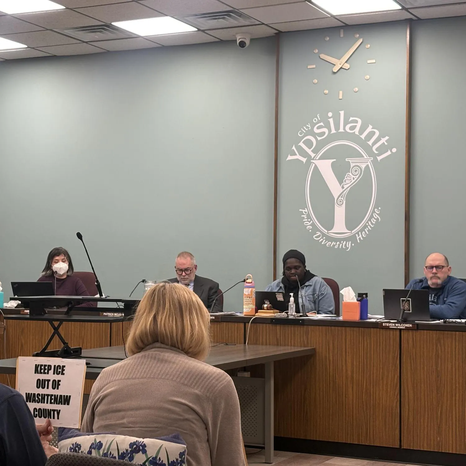 The back of two attendees in City Hall's Council Chambers watching on as councilors discuss a resolution.