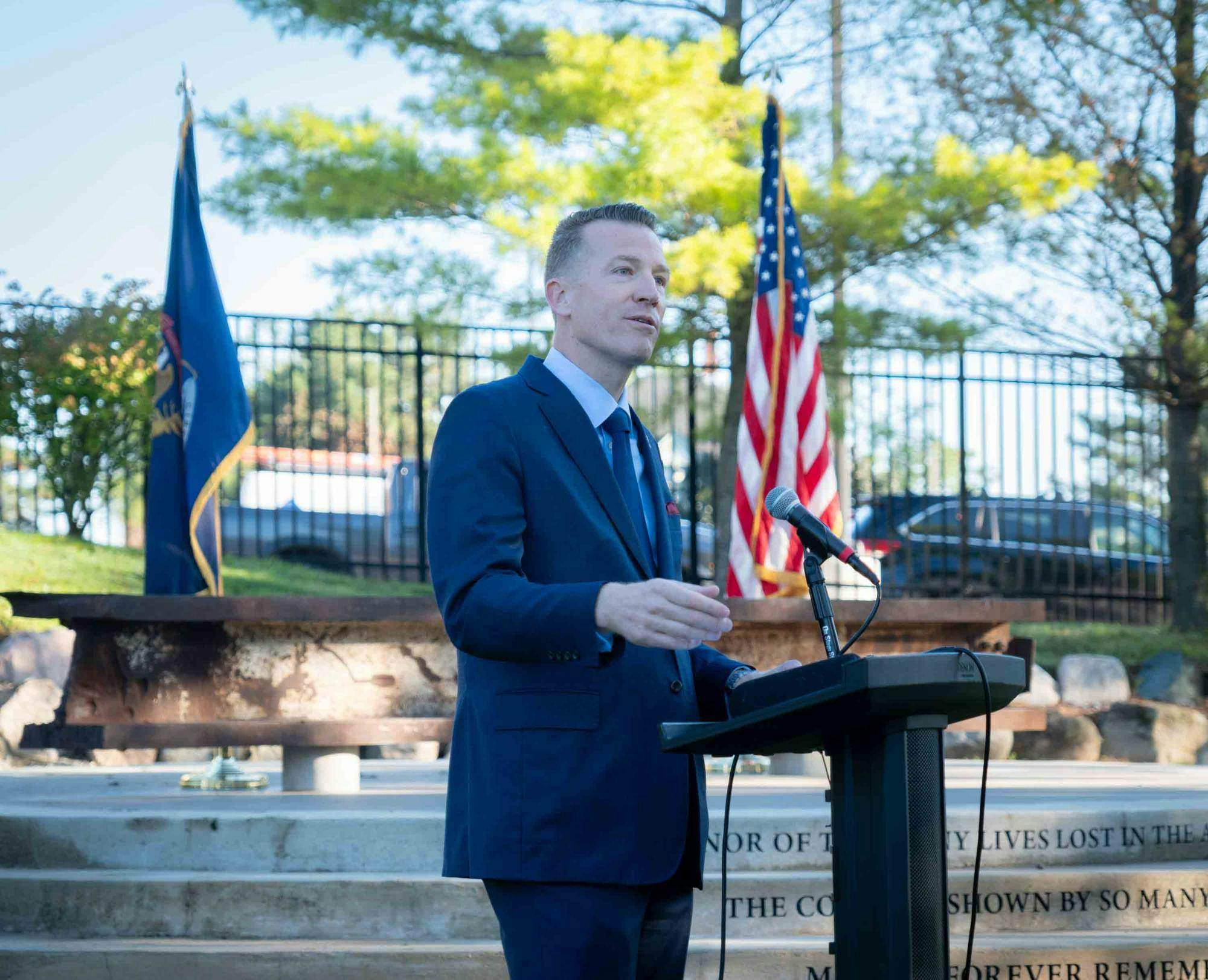 Brendan Kelly stands at a podium outside in front of a memorial on campus comprised of a steel beam from the South Tower of the World Trade Center. A Michigan and United States of America flag are both seen in the background. 